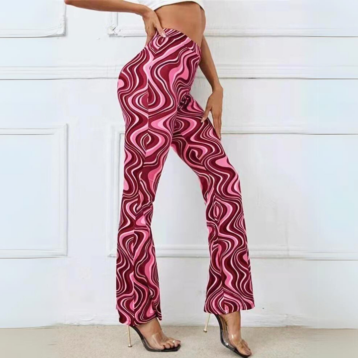 A person wearing trendy statement pants with a pink and maroon psychedelic swirl pattern, high-waisted and flared, paired with clear high-heeled shoes, poses against a white wall.
