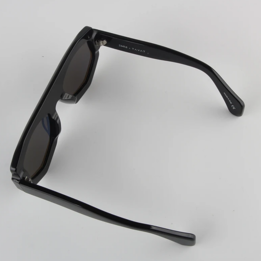 A pair of black geometric frame sunglasses with dark lenses, viewed from an angle above showing the inner arms and hinges, resting on a plain white surface. Crafted from acetate, they offer both style and UV400 protection.