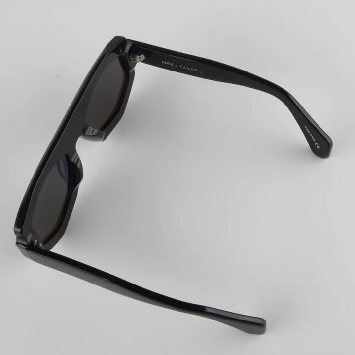 A pair of black geometric frame sunglasses with dark lenses, viewed from an angle above showing the inner arms and hinges, resting on a plain white surface. Crafted from acetate, they offer both style and UV400 protection.