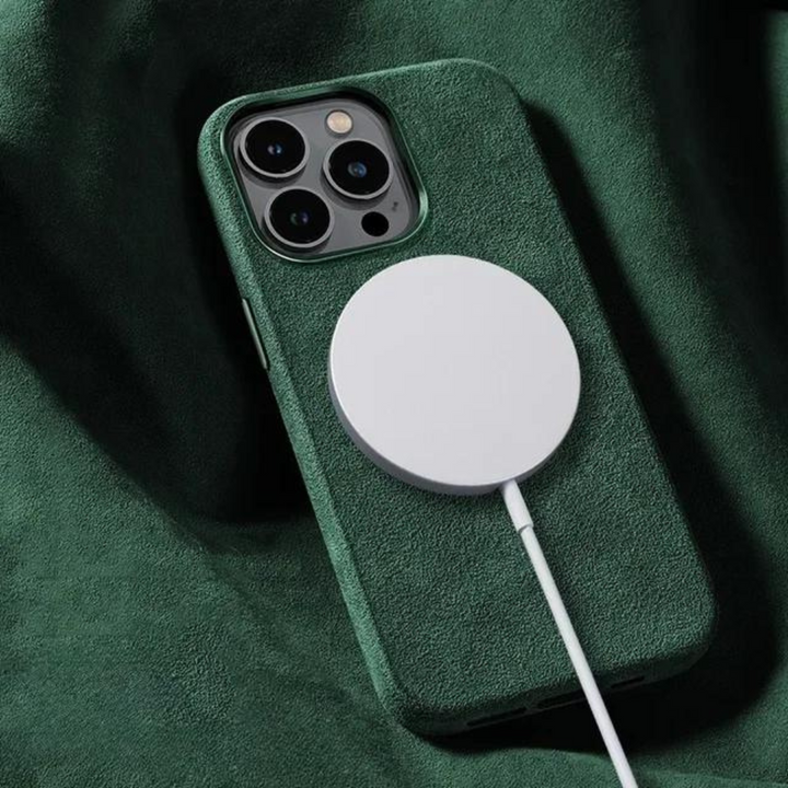 A smartphone with a green case featuring a suede finish is lying on a green fabric surface, with a circular wireless charger attached to its back and a charging cable connected.