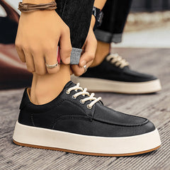 A person rolls up their jeans while wearing stylish black men's leather casual shoes with white laces and thick white soles, standing on a wood-patterned floor.