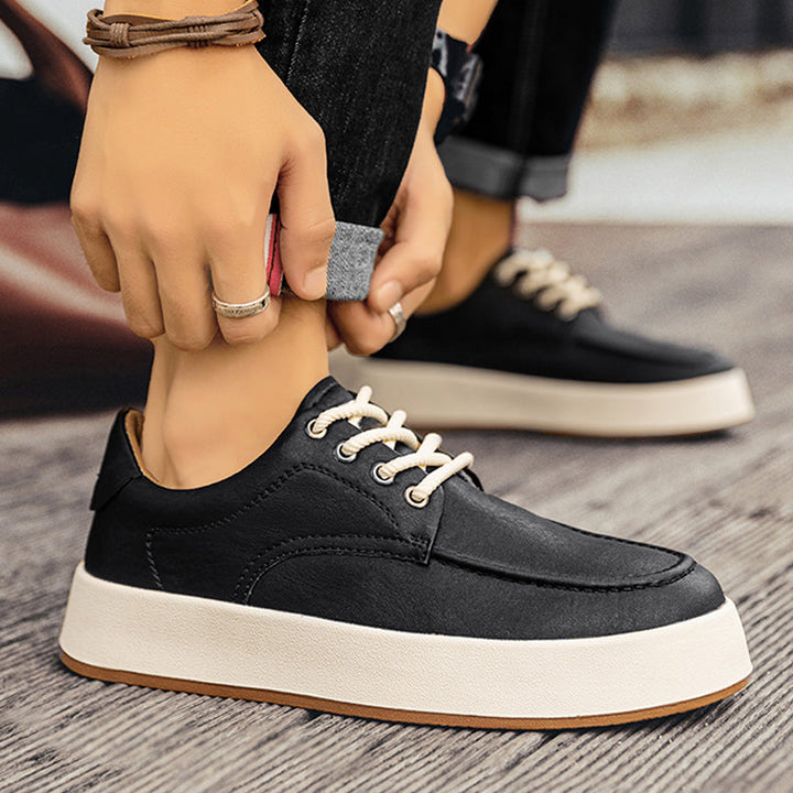 A person rolls up their jeans while wearing stylish black men's leather casual shoes with white laces and thick white soles, standing on a wood-patterned floor.