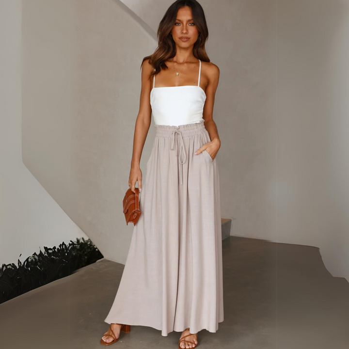 A woman in a white spaghetti strap top and flowy wide-leg beige pants with an elastic waistband stands indoors, holding a brown purse and wearing brown sandals. She has long, wavy hair and a relaxed, stylish look.