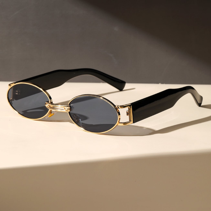 A pair of retro oval sunglasses with a durable metal frame, gold accents, and thick, glossy black arms rest on a light-colored surface against a dark background.