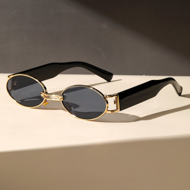 A pair of retro oval sunglasses with a durable metal frame, gold accents, and thick, glossy black arms rest on a light-colored surface against a dark background.