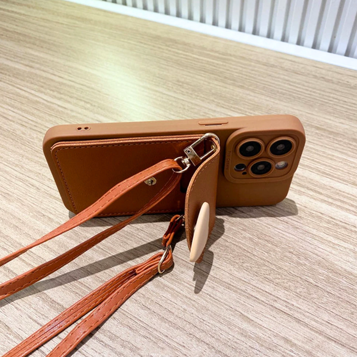 A brown smartphone with a chic phone case, featuring a detachable crossbody strap and a heart-shaped pocket, rests on a light wood table near a white slatted wall.