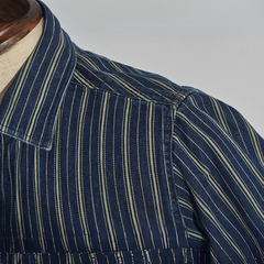 Close-up of a mannequin wearing a relaxed-fit button-down striped denim shirt in dark blue with vertical yellow and white stripes, featuring a pointed collar and visible shoulder and upper sleeve details.