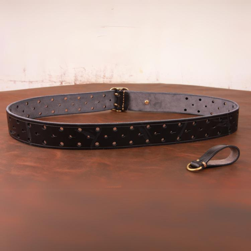 A black handcrafted belt with multiple holes and a gold buckle lies on a brown surface; a small matching leather loop with gold hardware is placed nearby.