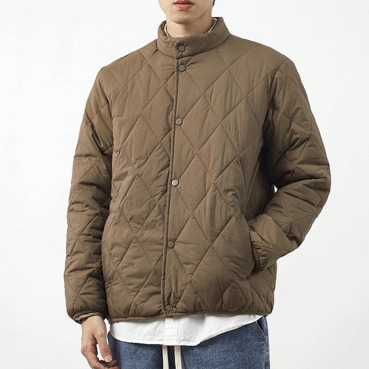 A person wearing a brown, diamond-stitched padded jacket with snap buttons, hands in pockets, over a white shirt and blue jeans against a plain white background.