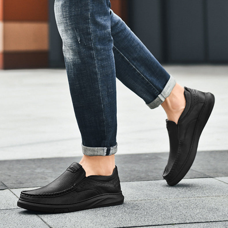 A person wearing rolled-up blue jeans and stylish men's cowhide leather slip-on shoes with a soft sole walks on a tiled pavement outdoors. Only the lower legs and feet are visible.