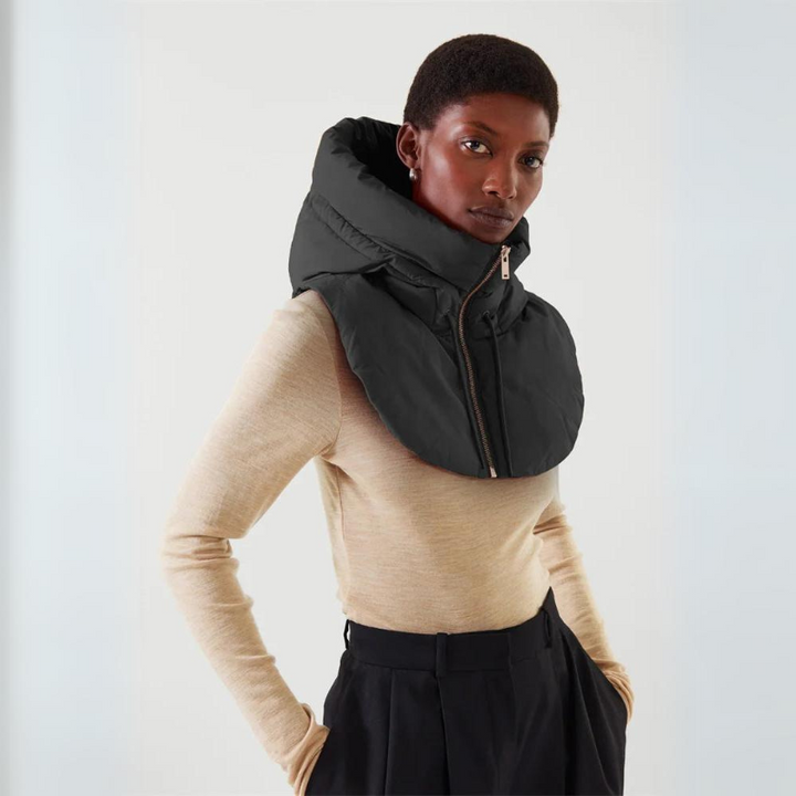 A person wearing a beige long-sleeve top, black high-waisted pants, and a lightweight, hooded design padded scarf with front zipper stands against a plain light background, looking confidently at the camera.