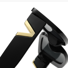 Close-up of the black temple arm of designer sunglasses, featuring a gold V-shaped accent. The lens and part of the geometric frame are partially visible in the background.
