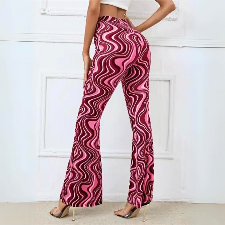 A person wearing trendy statement pants—high-waisted, pink and black swirl-patterned flared pants—and a white crop top stands indoors, posing with one hand on their hip and the other resting on a wall.