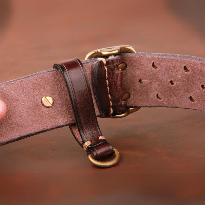Close-up of a handcrafted belt made from premium leather with a brass buckle and D-ring, featuring detailed stitching, multiple adjustment holes, and a suede-like underside against a brown background.