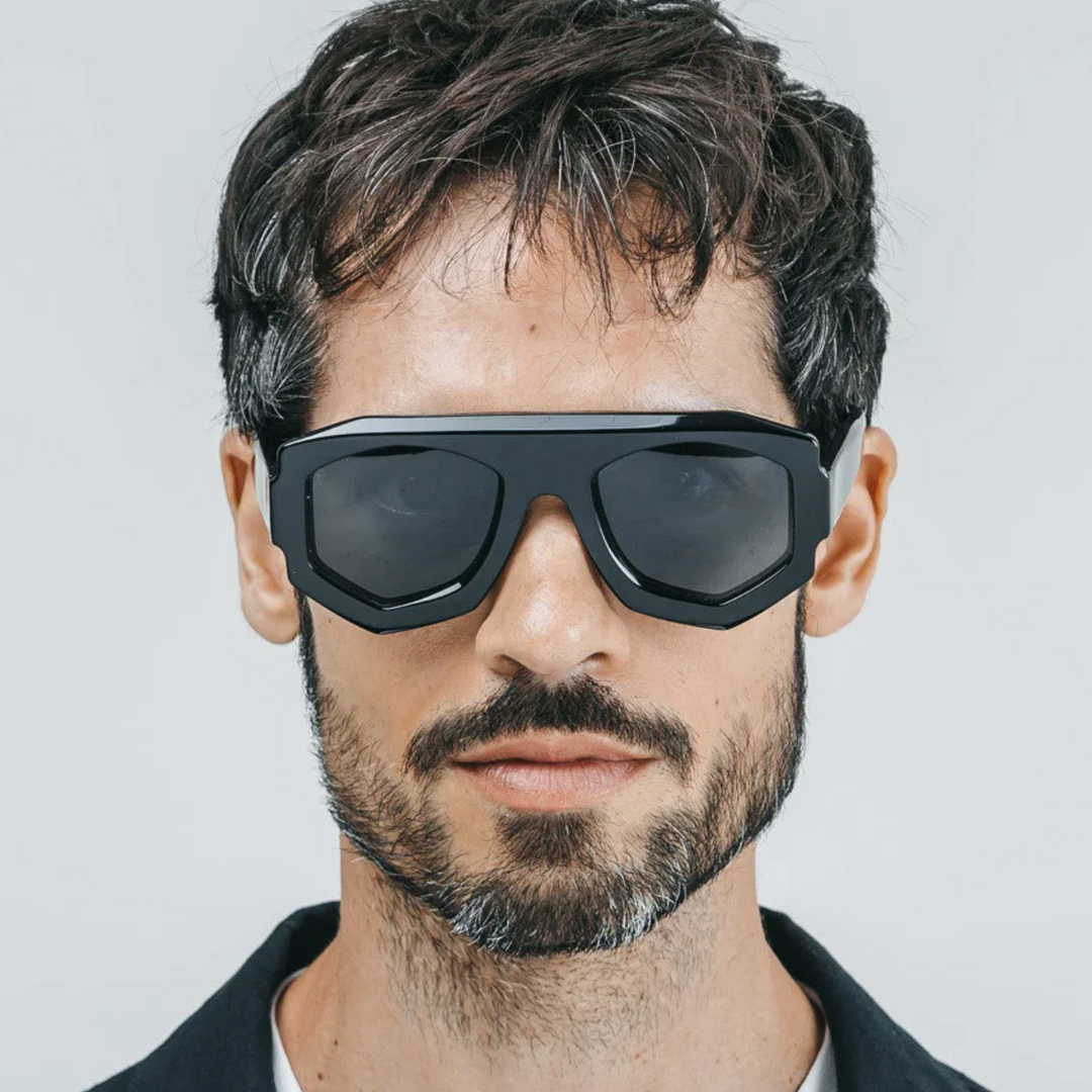 A man with short dark hair and a beard wears large, black acetate sunglasses with a geometric frame. He has a neutral expression and is facing forward against a plain light background.