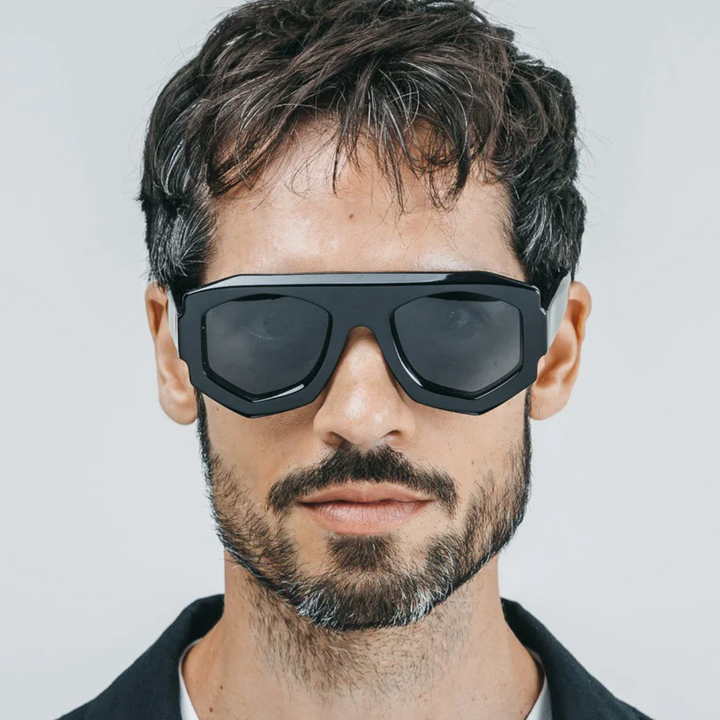 A man with short dark hair and a beard wears large, black acetate sunglasses with a geometric frame. He has a neutral expression and is facing forward against a plain light background.
