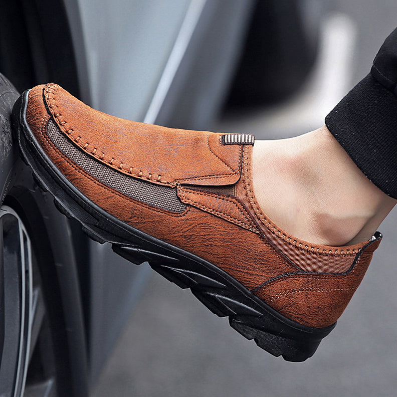 A close-up of a person wearing Men Leather Casual Shoes with black soles, resting their foot against a car tire. The shoe features visible stitching, a textured surface, and offers a comfortable fit for everyday wear.