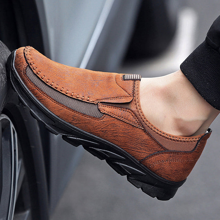 A close-up of a person wearing Men Leather Casual Shoes with black soles, resting their foot against a car tire. The shoe features visible stitching, a textured surface, and offers a comfortable fit for everyday wear.