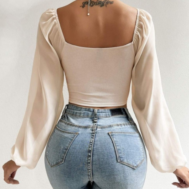 A woman is shown from the back wearing a cream-colored, long-sleeve crop top with a flirty neckline and high-waisted blue jeans. There is a tattoo on her upper back and a necklace hanging down her spine.