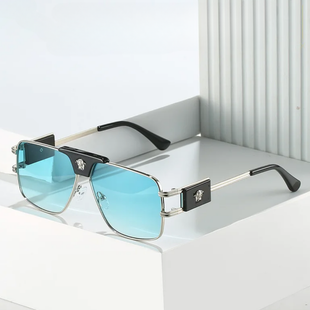 A pair of bold sunglasses with rectangular, blue-tinted lenses and premium metal frames, featuring black accents and a metallic emblem on the temples, displayed on a white geometric surface.