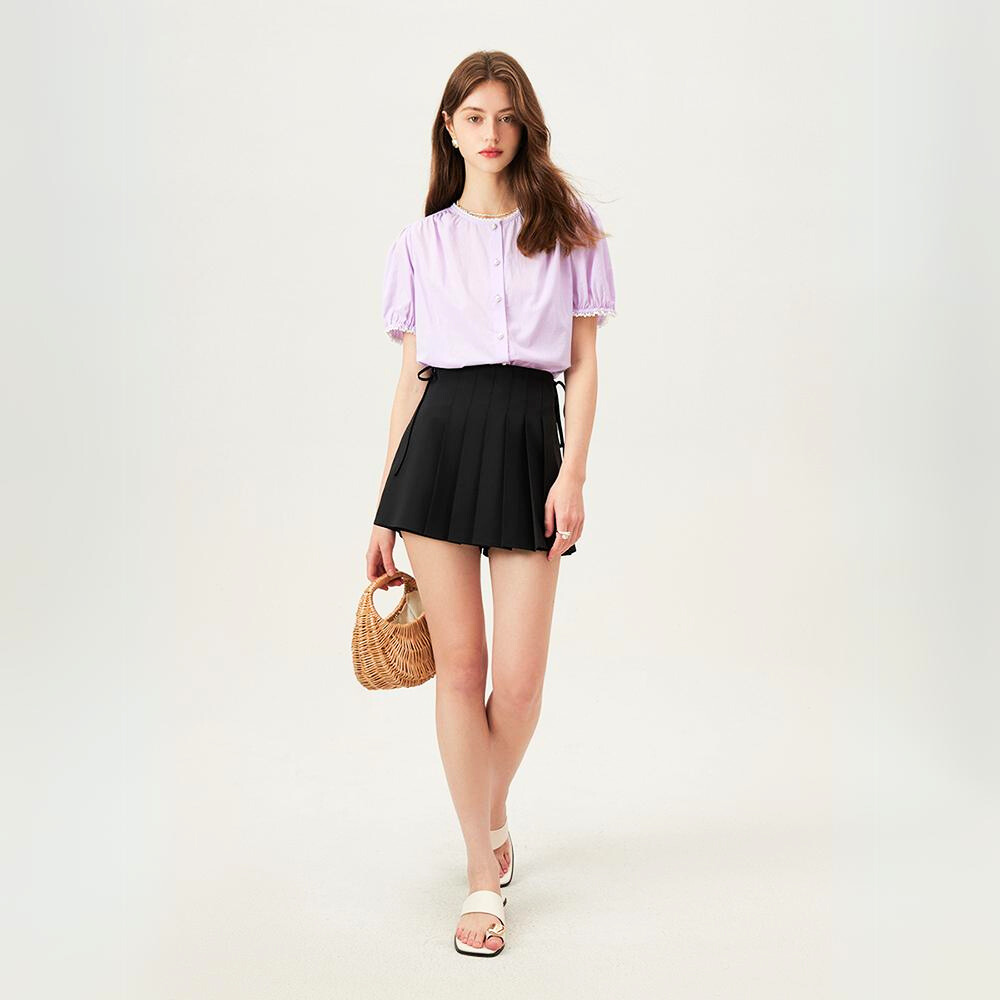 A woman stands against a plain light background, wearing a versatile blouse in light purple, a black pleated mini skirt, white sandals, and holding a small woven handbag. She has long brown hair and a relaxed expression.