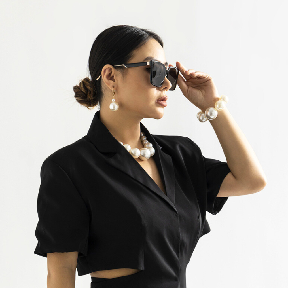 A woman wearing a black dress, oversized sunglasses, and a pearl jewelry set poses against a plain white background, her hair in a low bun as she adjusts her sunglasses with one hand.