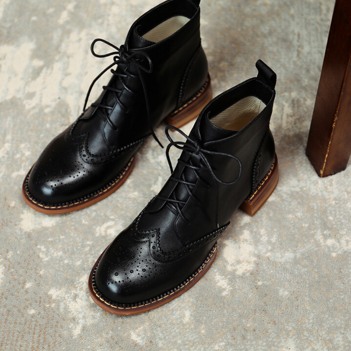 A pair of black leather brogue boots with decorative perforations and brown soles, crafted from genuine leather, placed on a light textured surface next to a wooden furniture leg.