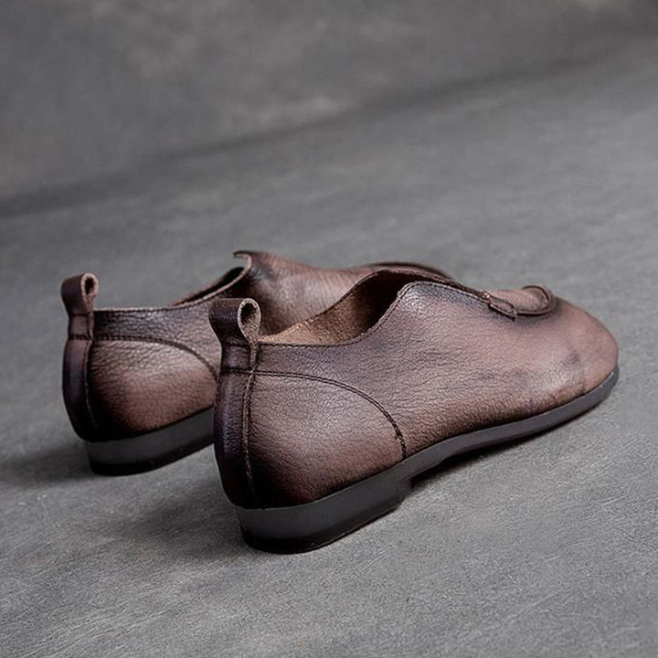 A pair of brown supple leather slip-on shoes with low black soles, placed on a gray textured surface, angled to show the back and side views.