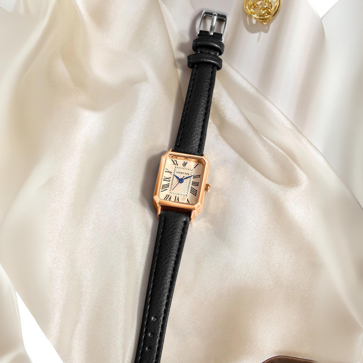 A rectangular face watch with a genuine leather strap and Roman numerals, displayed on smooth, cream-colored satin fabric near a gold decorative accessory.