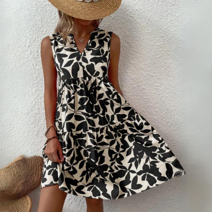 A woman wearing a sleeveless, black-and-white floral print dress crafted from lightweight breathable fabric and a wide-brimmed straw hat stands against a textured white wall, holding one side of her dress. Her face is partially hidden by the hat.