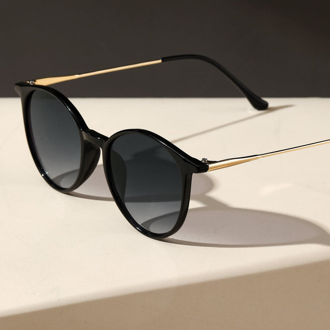 Black round sunglasses with gold arms rest on a light-colored surface, casting a soft shadow against a dark background—crafted for style and UV protection.