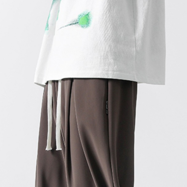 A person wearing a white shirt with green designs and loose brown GOAT Joggers with a drawstring waistband, shown from shoulders to knees against a plain background.