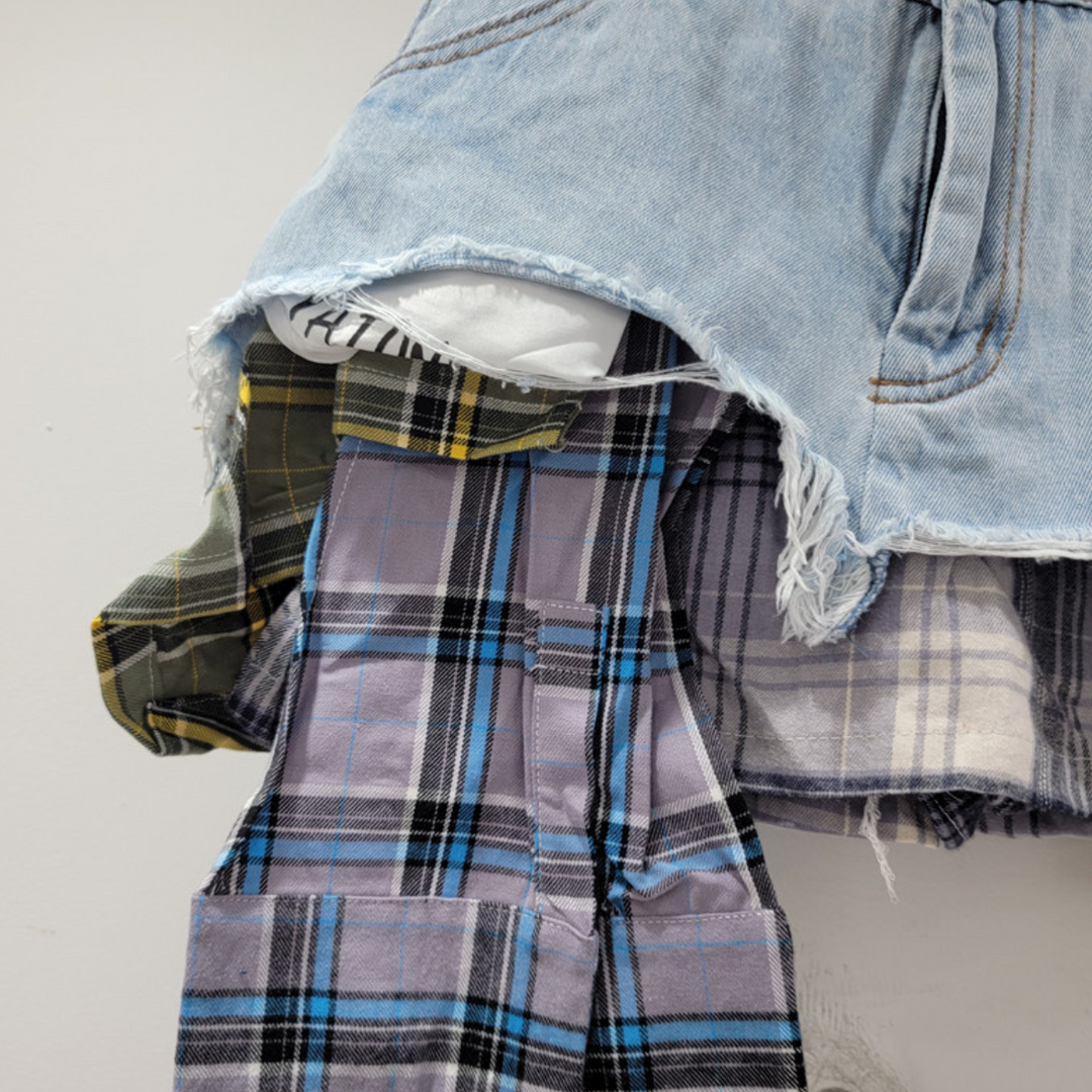 A close-up of a frayed denim and plaid patchwork skirt layered over yellow-black and purple plaid flannel shirts, hanging against a plain white background—perfect for showcasing eclectic fashion.