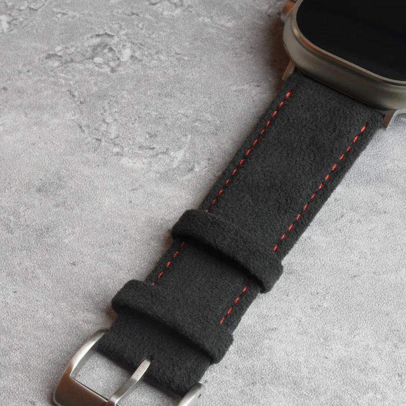 A close-up of a smartwatch with a black suede strap featuring orange stitching, a precision fit, and a stainless steel buckle with two loops, resting on a light grey textured surface.