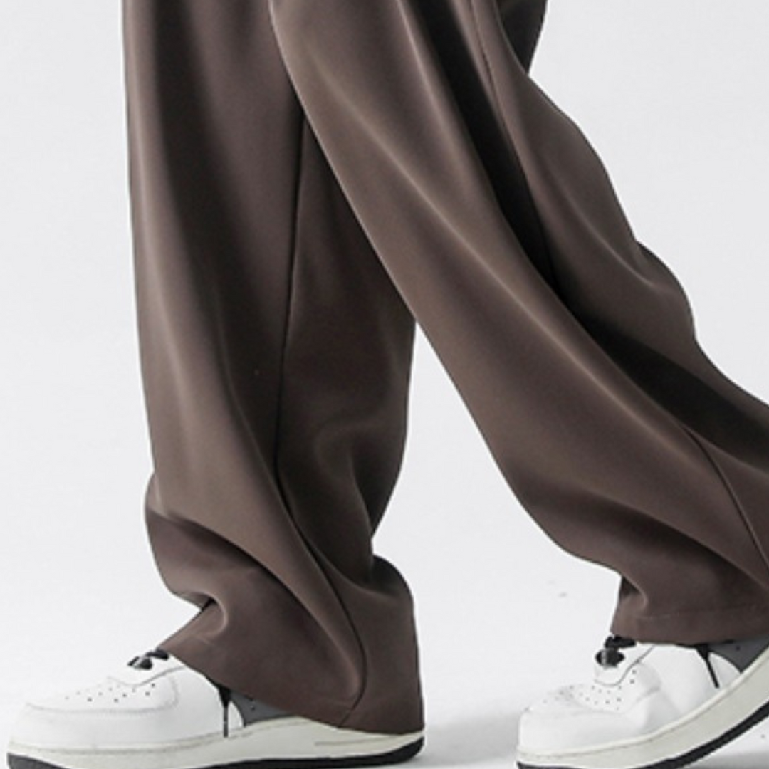 A close-up of a person wearing loose-fitting brown streetwear joggers and white sneakers with black soles, standing against a plain light background.
