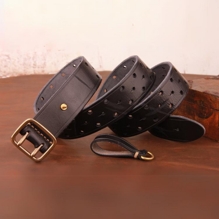 A handcrafted belt made from premium black leather with multiple holes and a brass buckle is coiled on a brown surface, accompanied by a matching leather keychain.