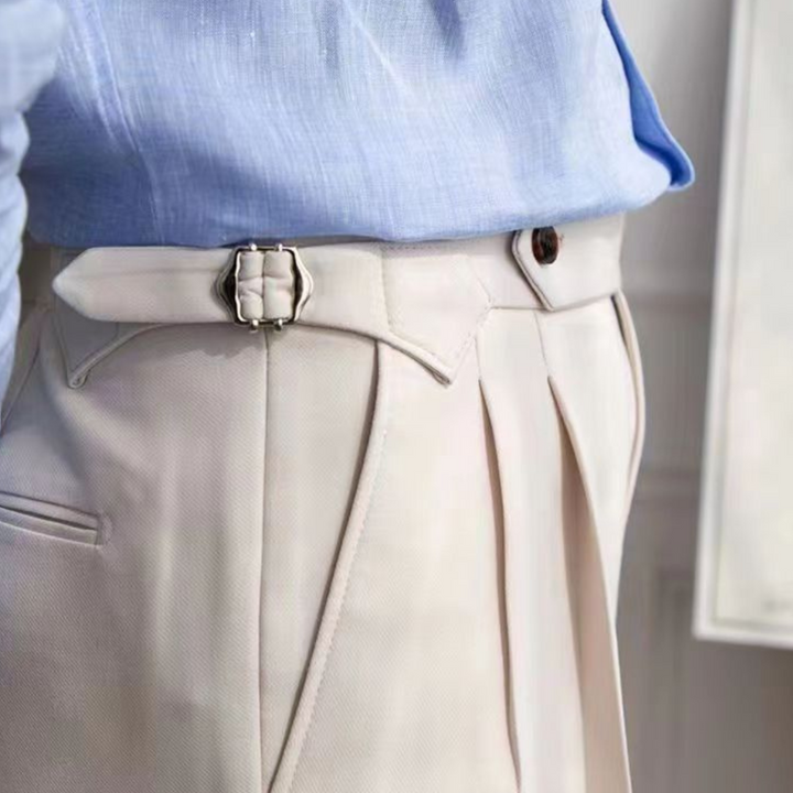 A close-up of a person wearing cream-colored, high-waisted trousers with a tailored fit and side buckle, styled with a tucked-in light blue shirt for an old money aesthetic.