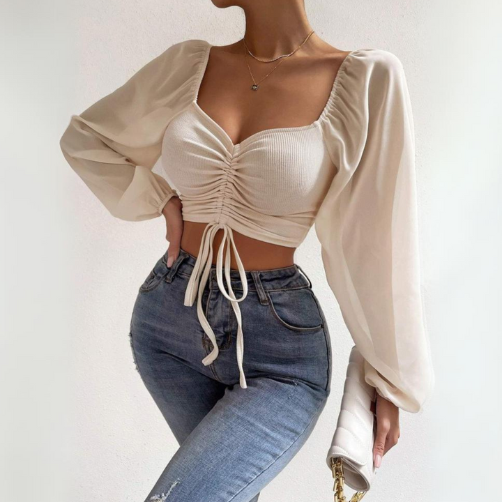 A woman wears a cream-colored, long-sleeve crop top with a ruched front and high-waisted blue jeans, posing against a plain background while holding a small white handbag. Her head is not visible in the image.