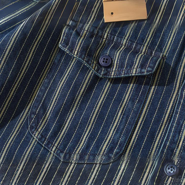 Close-up of a striped denim shirt with vertical white and yellow stripes, showing a front pocket with a button and part of a wooden hanger at the top.