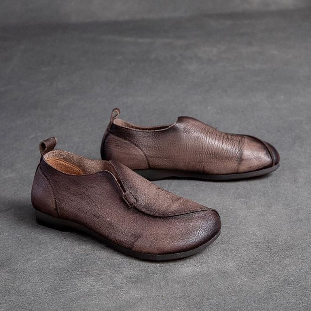 A pair of brown, minimalist leather slip-on shoes with rounded toes, low heels, and a cushioned insole, displayed on a gray textured surface.