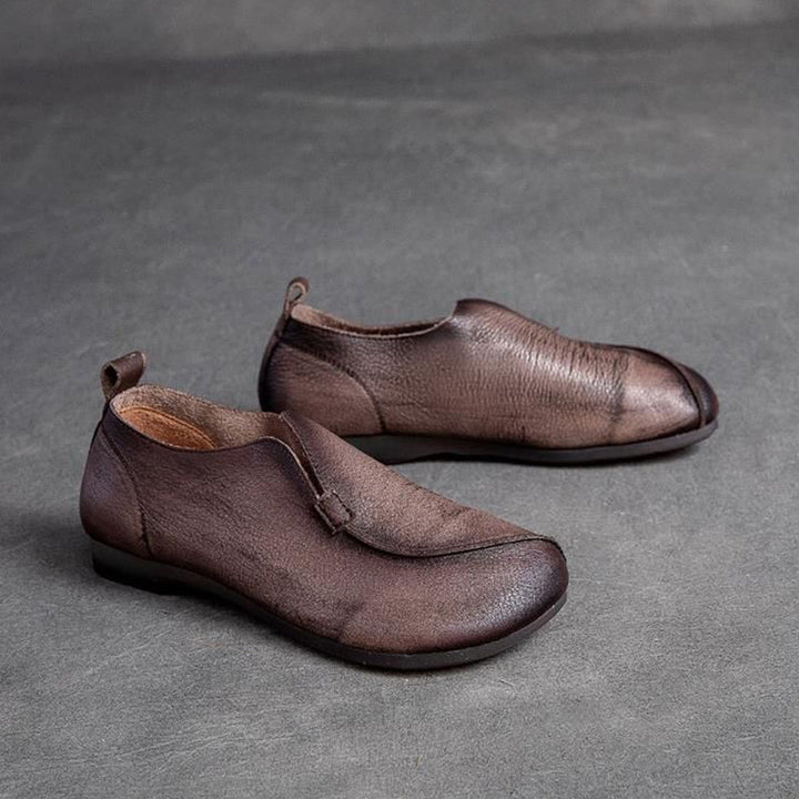 A pair of brown, minimalist leather slip-on shoes with rounded toes, low heels, and a cushioned insole, displayed on a gray textured surface.