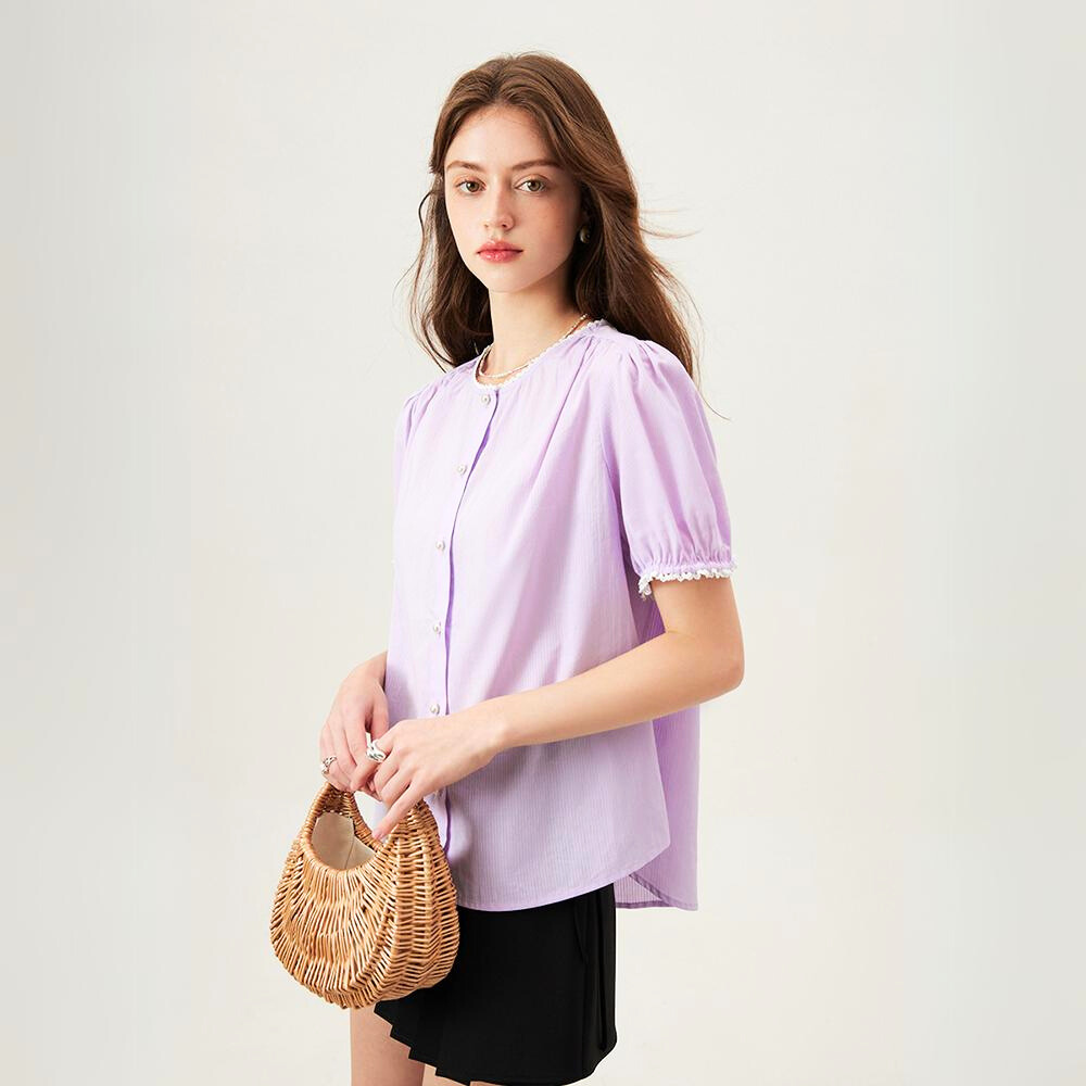 A woman with long brown hair wears a light purple, versatile blouse with short sleeves and black shorts, holding a round wicker handbag while standing against a plain light background.