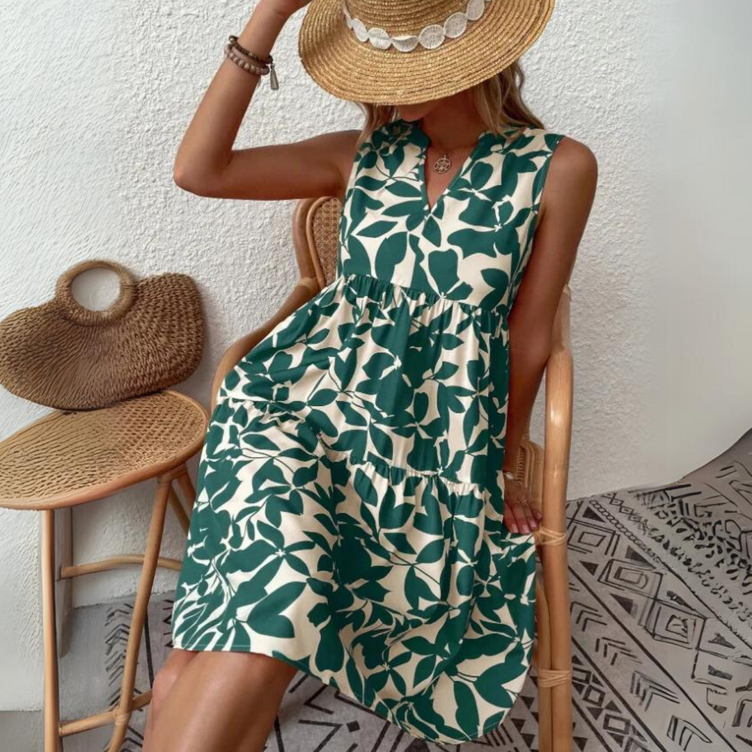 A woman in a leafy green and white sleeveless dress sits on a wooden chair, holding a wide-brimmed straw hat over her face. Crafted from lightweight breathable fabric, its the perfect addition to any summer wardrobe. A woven bag rests nearby on a patterned rug.
