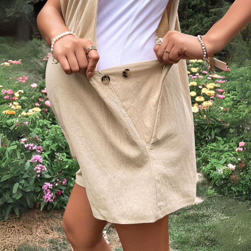 A person outdoors wears a beige wrap skirt and white top, holding the skirt’s buttoned flap open with both hands—an example of comfortable maternity clothes. Flower beds with colorful blooms and green foliage are visible in the background.