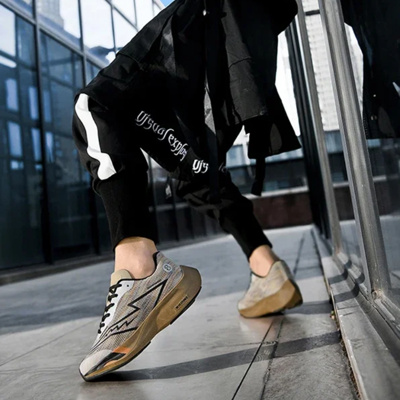 A person wearing black pants with a white stripe and beige sneakers featuring a shock-absorbing sole is walking on a city sidewalk next to a glass building. The low perspective emphasizes the comfort and style of the shoes.