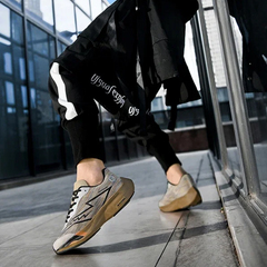 A person wearing black pants with a white stripe and beige sneakers featuring a shock-absorbing sole is walking on a city sidewalk next to a glass building. The low perspective emphasizes the comfort and style of the shoes.