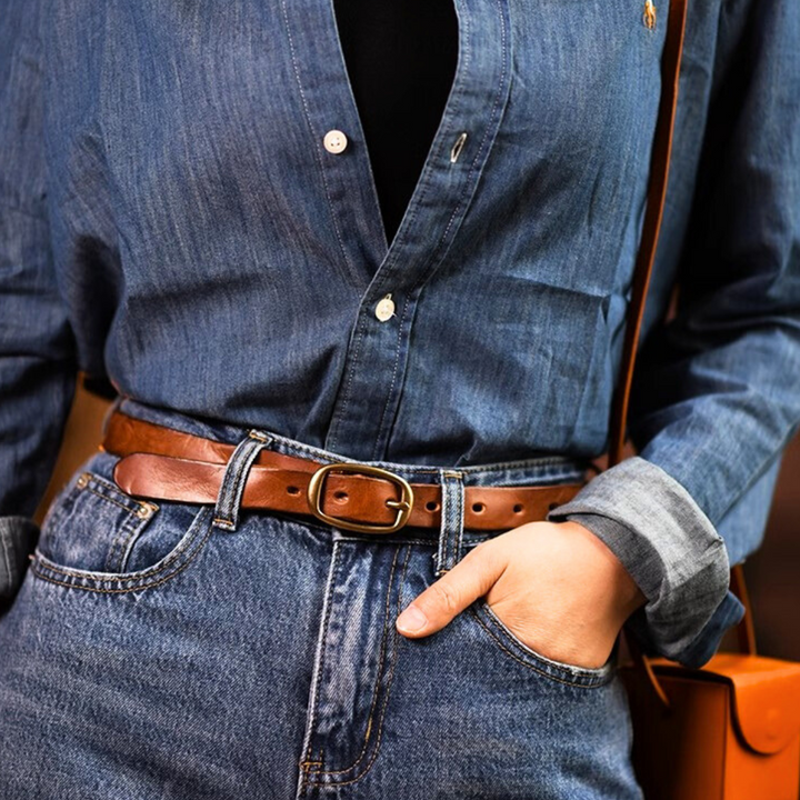 A person wearing a blue denim shirt tucked into high-waisted blue jeans with a leather belt, one hand in their pocket and a brown shoulder bag strap visible, showcasing classic country fashion.