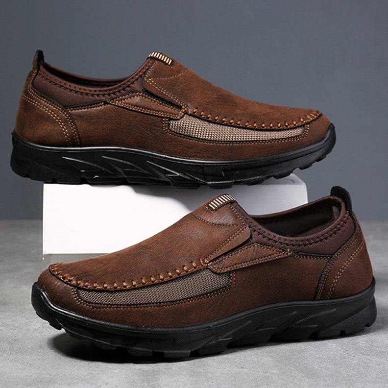 A pair of brown Men Leather Casual Shoes with black rubber soles and visible stitching, designed for a comfortable fit. Displayed against a gray background, one breathable loafer is propped up on a white block above the other.