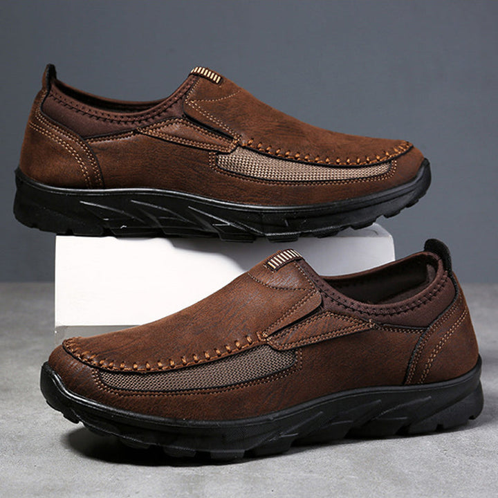 A pair of brown Men Leather Casual Shoes with black rubber soles and visible stitching, designed for a comfortable fit. Displayed against a gray background, one breathable loafer is propped up on a white block above the other.