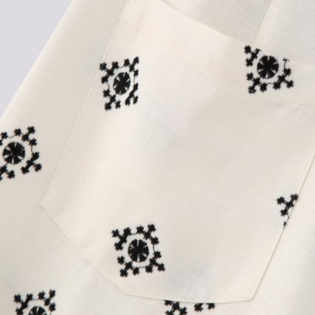 Close-up of a white linen fabric with a front pocket, featuring small black floral or star-like embroidered designs—perfect for pairing with wide-leg trousers or boho chic pants.